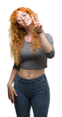 Wall Mural - Young redhead woman smiling looking to the camera showing fingers doing victory sign. Number two.