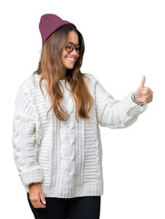 Wall Mural - Young beautiful brunette hipster woman wearing glasses and winter hat over isolated background Looking proud, smiling doing thumbs up gesture to the side