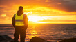 © Kiron - A solitary figure in reflective safety gear stands by the ocean, gazing at the golden sunset, capturing a serene moment of reflection and inner calm. Ai generated