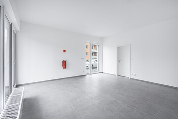 Wall Mural - Bright, empty room with gray tiled floor, white walls, and large windows, featuring a door leading outside. A fire extinguisher is mounted on the wall, adding a safety element to the modern space.