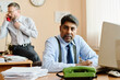 © pressmaster - Portrait of businessman sitting at desk, focusing on work while colleague talks on phone in background, office environment with various documents and devices present