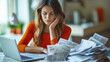 © Maksym - A stressed woman sitting at her desk, reviewing her expenses on a laptop, with bills piling up beside her