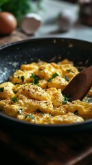 Yellow scrambled eggs in black frying pan, seasoned with herbs and pepper, wooden spatula