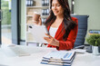 © NanSan - An Asian businesswoman sits at her desk, working efficiently on a computer and tablet. She smiles confidently, displaying her expertise as a financial analyst. Her work brings her happiness