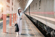 © Satori Studio - A young woman traveler with a backpack and camera stands on a train station platform, ready for her journey.