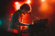 © VisualProduction - A musician performing on stage, playing a keyboard with a microphone. The scene is illuminated by vibrant red and orange lights, creating a dramatic atmosphere.