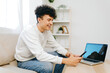 © SHOTPRIME STUDIO - Young man smiling while using smartphone and sitting on a couch next to a laptop in a bright, modern living space Light colors and greenery create an inviting atmosphere