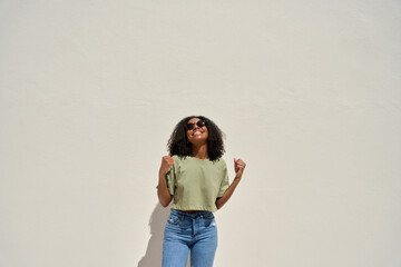 Wall Mural - Happy young African American woman winner feeling joy celebrating win victory standing at white wall background. Funky gen z Afro Black girl dancing having fun at sunny summer party. Copy space.