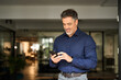 © insta_photos - Smiling mature business man corporate leader using cellphone standing in office. Happy busy middle aged older male entrepreneur holding mobile phone tech device looking at smartphone at work.
