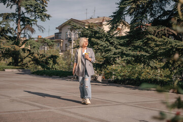 Wall Mural - A woman is walking down a sidewalk with a cup in her hand. She is wearing a jacket and jeans.