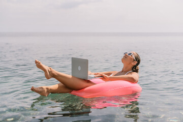 Wall Mural - A woman is floating on a pink inflatable raft in the ocean while holding a laptop. Concept of relaxation and leisure, as the woman is enjoying her time in the water while working on her laptop.