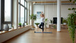 © khonkangrua - A woman performs a yoga pose in a spacious, light-filled studio, surrounded by indoor plants and large windows.