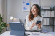 © amnaj - Young asian businesswoman is sitting at her desk in her office, smiling and using her smartphone