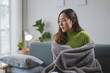 © amnaj - Young asian woman feeling sick and having cold, covering herself with a blanket, while sitting on a sofa at home