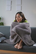 © amnaj - Young woman sits on a cozy sofa at home, wrapped in a warm blanket, battling the winter chill and feeling under the weather. She looks unhappy and worried, lost in thought, barefoot and casual