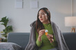 © amnaj - Sick young woman is sitting on her sofa wrapped in a blanket and holding a pill bottle