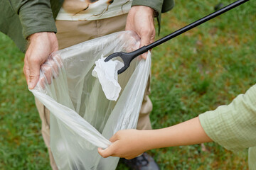 Two people are using a grabber tool to pick up trash and place it in a plastic bag while standing on grass. They are engaged in a clean-up activity without wearing gloves