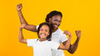 © Prostock-studio - Celebrating Success. Cheerful black mother and daughter holding hands up for victory isolated on yellow background