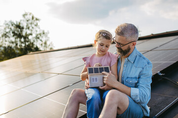 Daughter and father on roof with solar panels, holding model of house with rooftop photovoltaic system. Sustainable future for next generation.