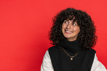 Portrait of happy Caucasian young woman with curly hair smiling friendly, glad expression looking at camera dreaming, resting, relaxation feel satisfied good news. Girl indoors on red background