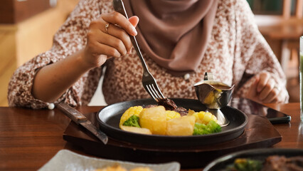 a woman wearing a hijab is holding a fork and knife to cut beef steak on a hotplate