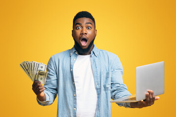 Dazed afro man with dollar cash and laptop over yellow studio background with free space