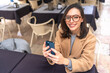 © Andrii - Happy middle-aged woman using smartphone sitting outdoor cafe. Smile businesswoman in glasses dressed trench coat holding smartphone in hand looking at camera. High angle view. Copy space