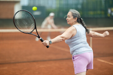 Wall Mural - Rear view of emotional aged woman enthusiastically playing tennis outdoors in summer, preparing to hit forehand to return ball to female opponent court during friendly match