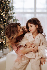 A woman is holding a young girl in a pink pajama outfit. The girl is smiling and the woman is looking at her. The scene is set in a room with a Christmas tree in the background