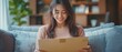 © Sawitree88 - A young woman, seated comfortably at home, smiles warmly as she unboxes a package.