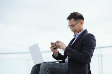 A confident Asian businessman standing in a modern city, smiling while holding a smartphone, dressed in a sharp suit and glasses, embodying corporate success and professionalism.