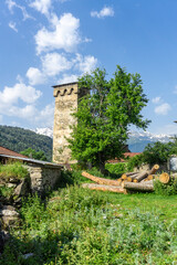 Wall Mural - Medieval defensive stone tower with loopholes near the house. Trees, green grass, sawn tree logs and bushes. Hills covered with bushes, mountains and snow