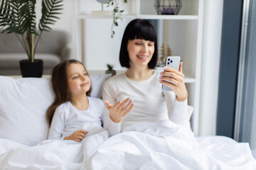 Wall Mural - Mom and daughter lying in bed having video call talking on phone. They appear happy and relaxed, enjoying morning together.