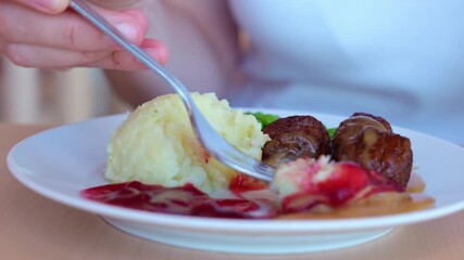 Wall Mural - Fork lifting a swedish meatballs from plate with mashed potatoes, gravy and lingonberry sauce. Woman eating traditional meal in public food court