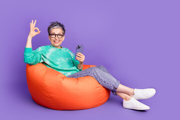 Sticker - Full body photo of attractive senior woman sit beanbag hold device okey dressed stylish green clothes isolated on purple color background