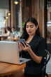 © bongkarn - An attractive Asian businesswoman is using her smartphone while working remotely from a cafe.