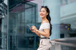 © bongkarn - An elegant Asian woman with a smartphone in her hand is standing in front of the building.