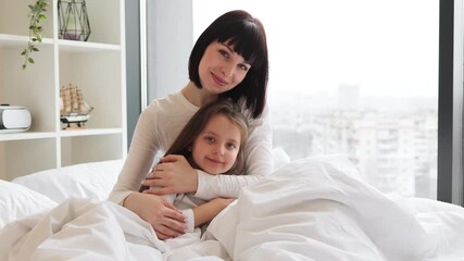 Wall Mural - Mother and daughter hugging in bed in bright, cozy bedroom. They look content and happy, sharing a moment of love and comfort. Bonding between parent and child in a serene, peaceful setting.