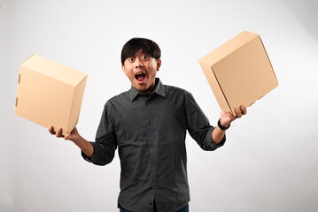 Asian man in a dark gray shirt holding two cardboard boxes, one in each hand, against a white background. The man appears cheerful, possibly promoting packaging, shipping, or gift services.