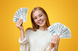 © Prostock-studio - Young investor. Happy millennial girl holding lots of dollars on orange background with free space