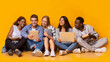 © Prostock-studio - Group of multiracial students studying for university exams, sitting on floor, using laptop, digital tablet, holding books, yellow background