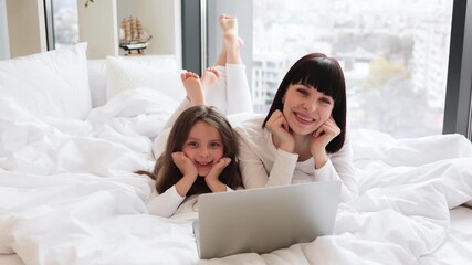 Wall Mural - Mother and daughter lying on bed using laptop together. Happy family bonding in cozy home environment. Smiling faces show love, togetherness, and relaxation.