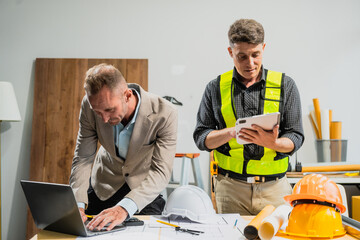 Wall Mural - A Caucasian middle-aged male engineer contractor and an Italian architect engineer are seated at a desk, discussing renovation plans with blueprints and a house model in front of them.