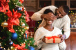 © Prostock-studio - Christmas gift. African american man surprising wife with xmas gift, closing her eyes and giving present in decorated living room