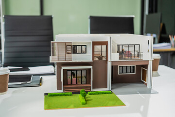 Wall Mural - A modern engineer's desk featuring a detailed house model, blueprints, and renovation plans. The wooden desk is neatly organized in a bright, glass-walled room, with no people present.