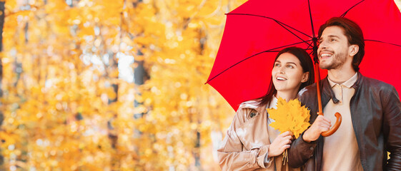 Happy lovers walking under red umbrella at rainy autumn day by forest, panorama with free space