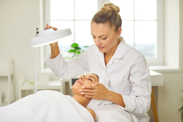 Wall Mural - Young professional woman cosmetologist doing skin care facial treatment examining its condition for female client with lamp in beauty clinic. Cosmetology and beauty procedure concept.