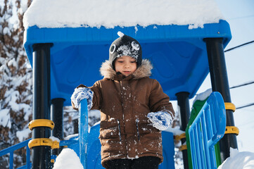 A toddler in a brown jacket and black hat is happily playing in the freezing snow in a public space, enjoying leisure fun in the winter.