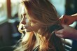 © Victor Bertrand - A woman with long wavy blond hair getting her hair gently brushed by a stylist, bathed in soft natural light, creating a serene atmosphere.