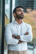 © Andrii Zastrozhnov - A man in a white shirt and tan pants is standing in front of a window, looking out at the street. He is wearing a watch and has his arms crossed. Concept of contemplation and introspection